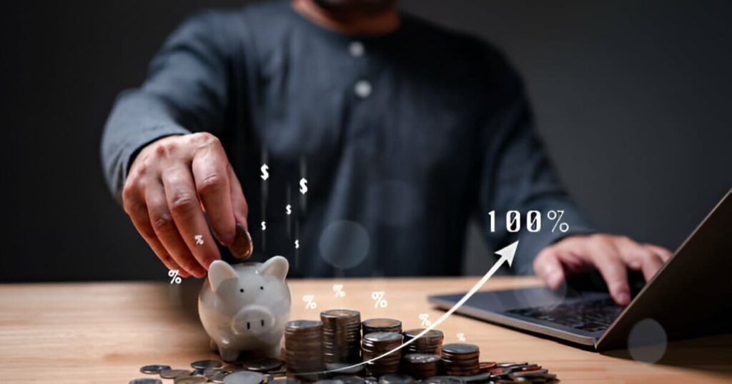 Hombre frente a una laptop y pilas de monedas, guardando una en una alcancía
