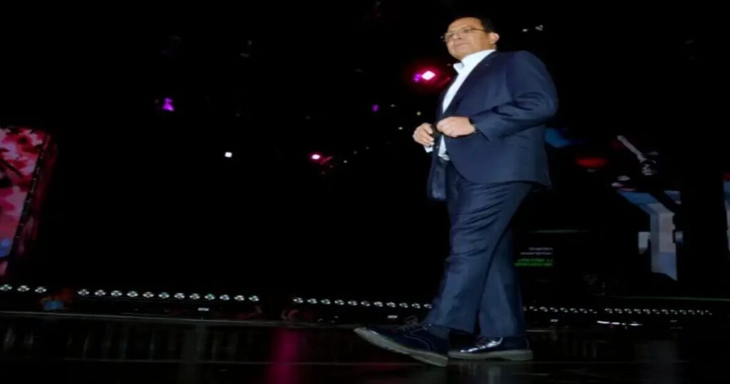Ernesto Reséndiz con traje azul y camisa blanca caminando en un escenario iluminado con pantallas de fondo durante un evento corporativo
