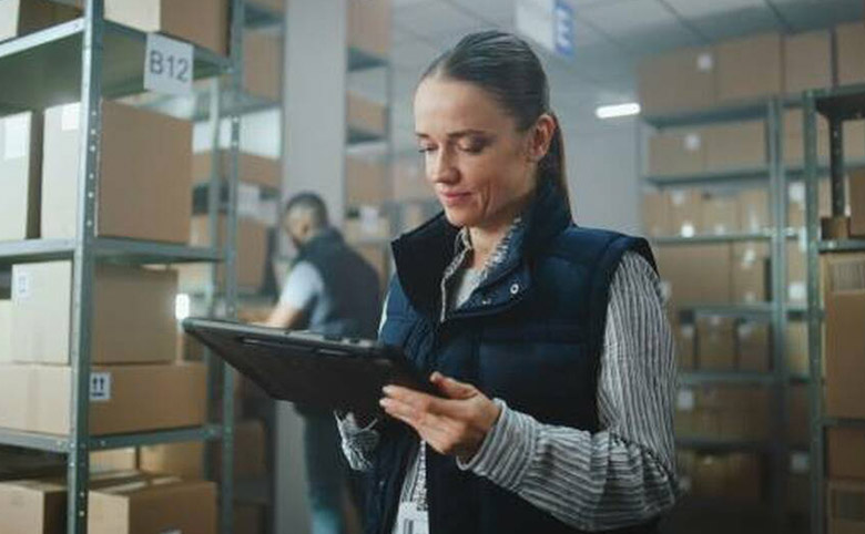 Mujer entusiasta mirando una tableta electrónica dentro de un almacén con una persona acomodando una caja al fondo