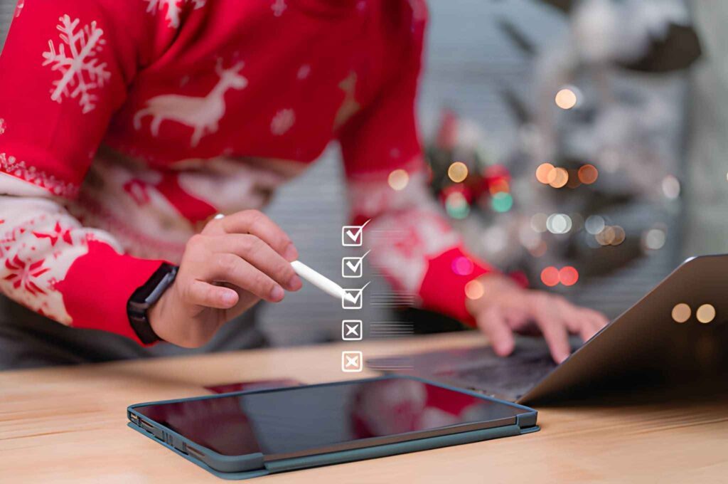 Persona con suéter navideño, lápiz óptico, tableta y laptop realizando checklist virtual 